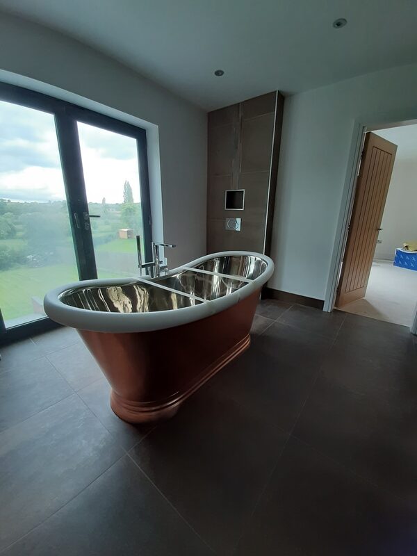 Copper roll-top bath standing on a dark tiled floor in front of a full-height window looking out over fields