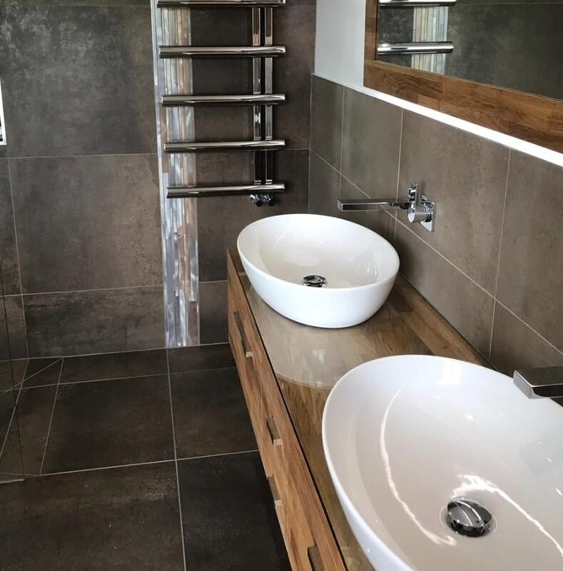 Two white countertop basins on an oak vanity against a dark porcelain wall, with a chrome ladder radiator behind
