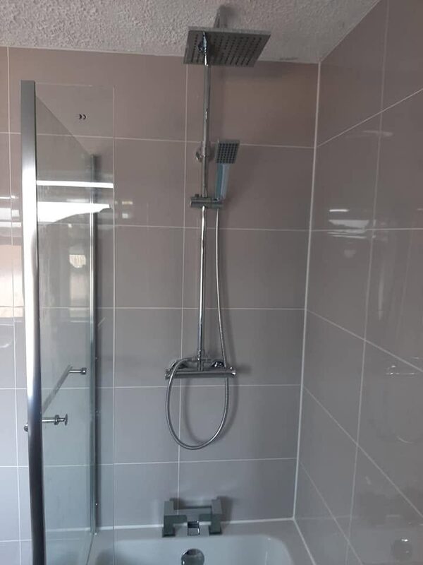 Square rainfall shower head and riser over a bath, against pale grey gloss tiles behind a glass screen