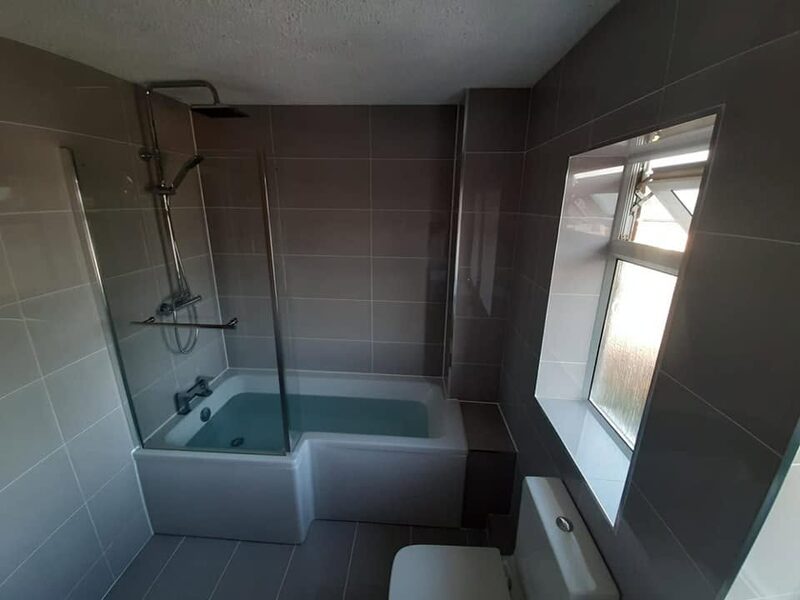 L-shaped bath with a curved glass screen in a bathroom tiled in dark grey, with a window to the right