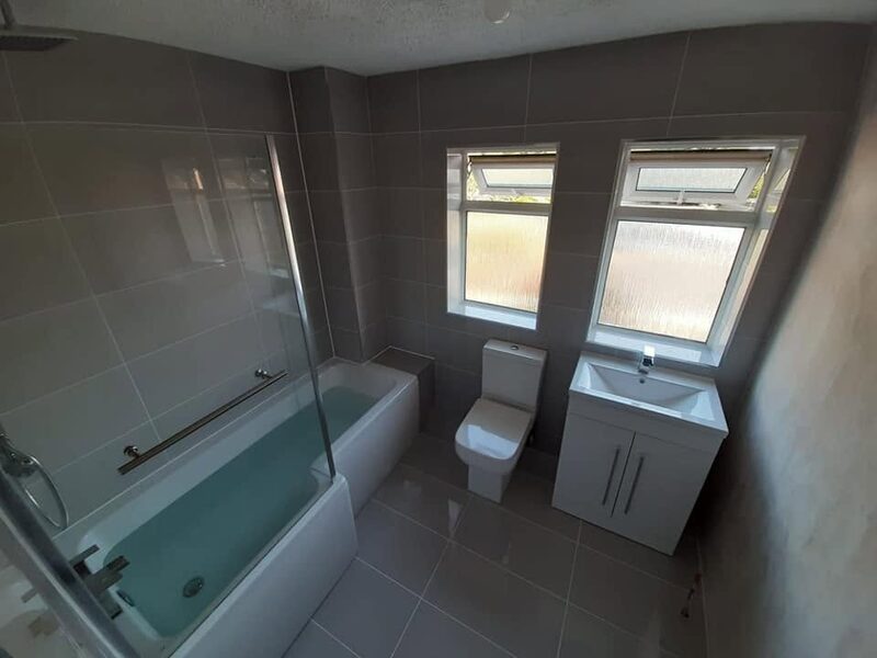 Grey tiled bathroom under a pitched ceiling, with a bath and glass screen on the left and two windows straight ahead